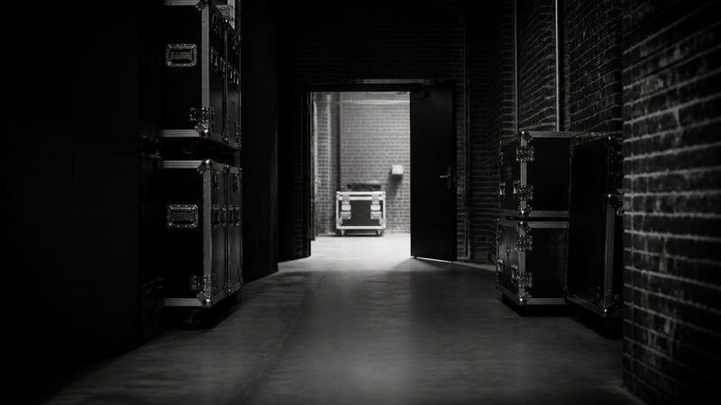 Empty backstage corridor of a concert venue, flight cases stacked against brick walls, a single door propped open