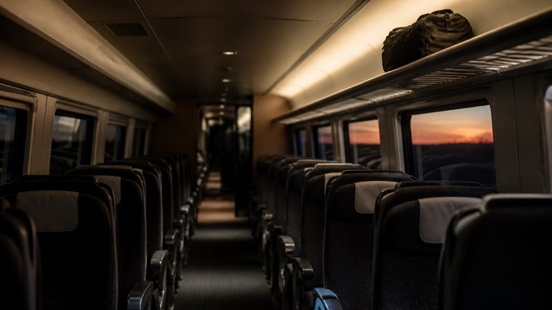 Empty high-speed train carriage at dusk, rows of vacant seats, a single overhead luggage rack holding one abandoned bag