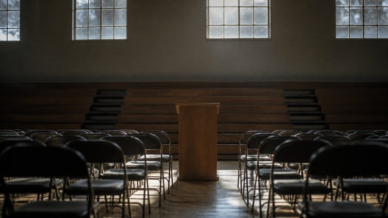 Rows of empty folding chairs facing a bare wooden podium in a provincial gymnasium, morning light through high windows