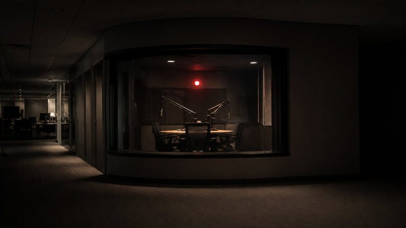 Late-night newsroom corridor with an empty radio broadcast booth behind glass, red light glowing, chairs pushed back from microphones